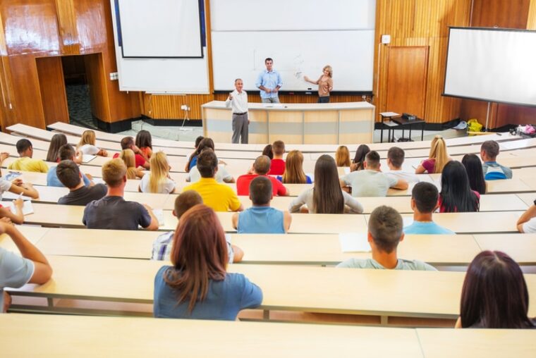 Ein halbvoller Hörsaal voller Studierenden, die drei Professor*innen zuhören.