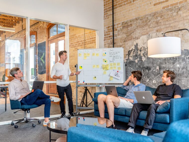 Drei Männer sitzen im Büro und hören einen anderen Mann zu wie er redet und etwas an einem Whiteboard mit Sticky Notes erklärt