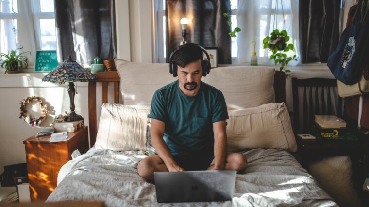 Mann sitzt mit Kopfhörern im Bett und schreibt etwas am Laptop