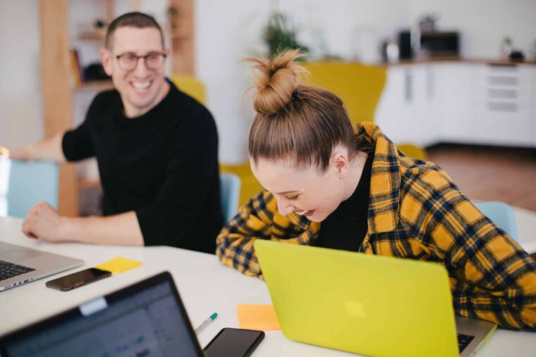 Zwei Kollegen im Büro sitzen am Tisch vor ihren Laptops und lachen