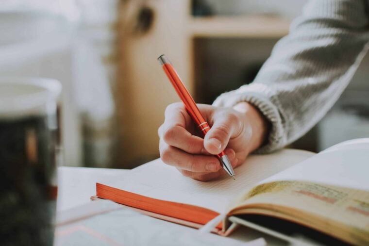 Person sitz am Schreibtisch und schreibt mit einem roten Stift in ein Notizbuch