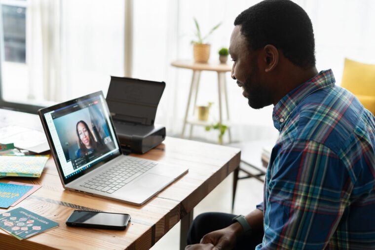 Mann sitzt am Tisch vor seinem Laptop und spricht mit seinem Kollegen der auf dem Bildschirm zu sehen ist