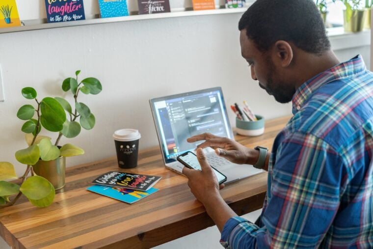 Ein Mann sitzt am Schreibtisch vor seinem Laptop und tippt etwas auf sein Handy