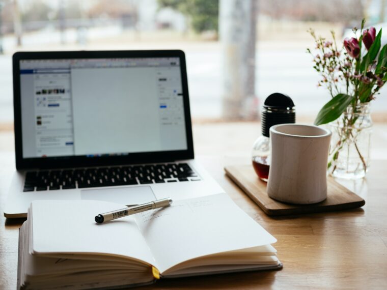 Ein Laptop steht auf einem Schreibtisch, ein Notizbuch liegt davor und eine Tasse rechts daneben