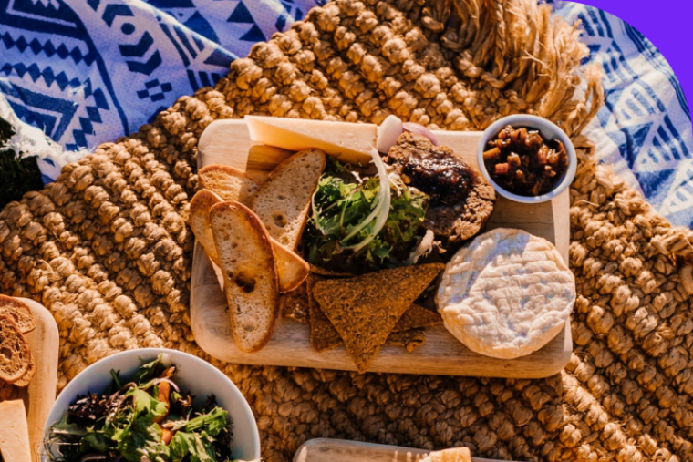 Ein Frühstücksbrett mit geschnittenem Brot, Salat, und Brie auf einer Picknickdecke.