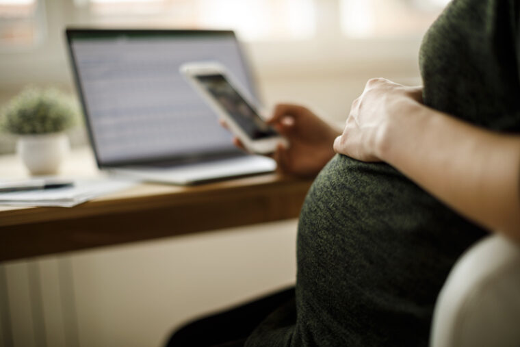 Eine schwangere Frau schaut sich während der Arbeit am Laptop ihr Handy an.