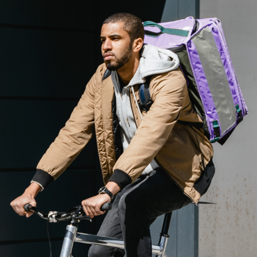 Mit großem Rucksack ausgestattet fährt ein junger Mann im Rahmen seines Nebenjobs auf dem Fahrrad durch die Stadt, um Lebensmittel an die Kundschaft zu liefern.