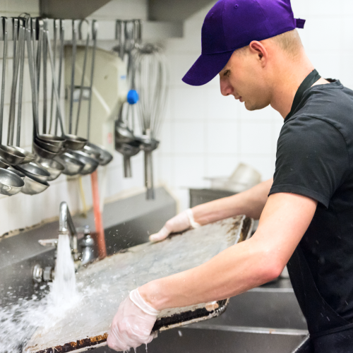 One of the most popular summer jobs in our app is working as a kitchen helper. The image shows a young man washing a large pan in a restaurant kitchen.
