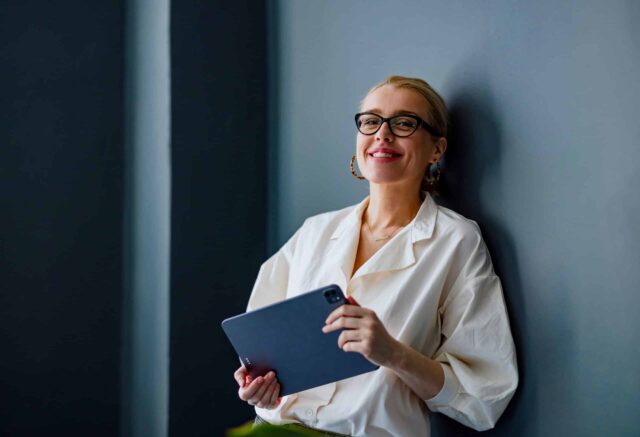 A woman holding a tablet is leaning against a wall
