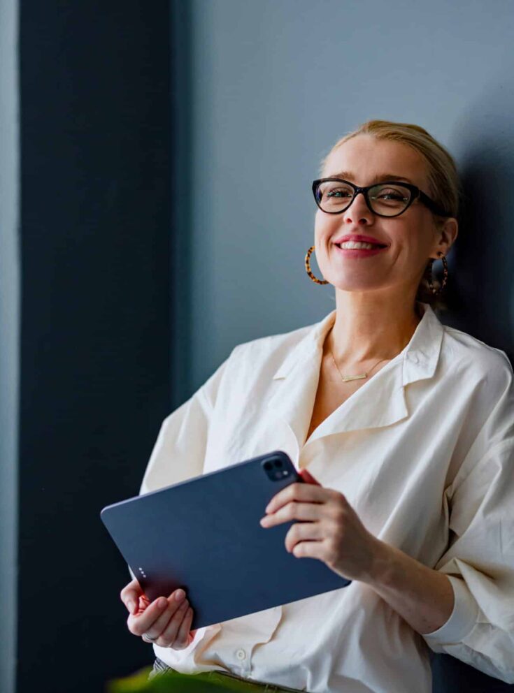 A woman holding a tablet is leaning against a wall